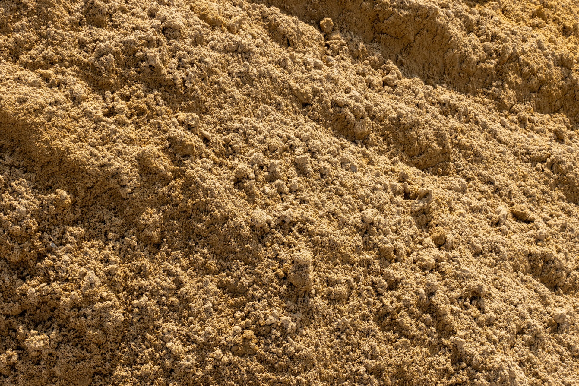 A large mound of coarse, brown sand fills the frame, with loose texture and uneven surface, under bright light revealing fine grains.