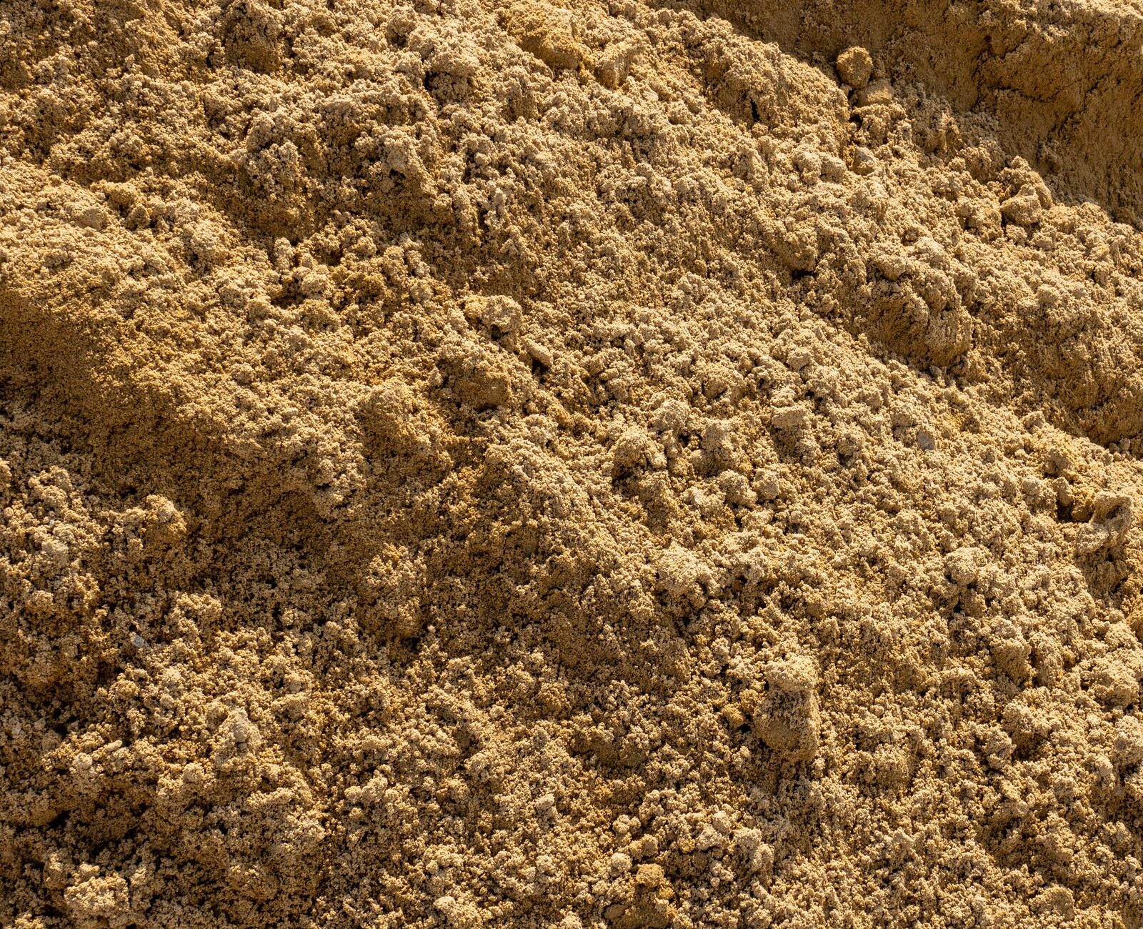A large mound of coarse, brown sand fills the frame, with loose texture and uneven surface, under bright light revealing fine grains.