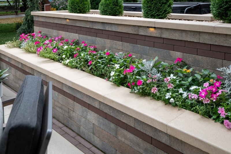 A stone planter box with vibrant pink and white flowers set against a neutral-toned brick wall, surrounded by greenery and patio furniture.