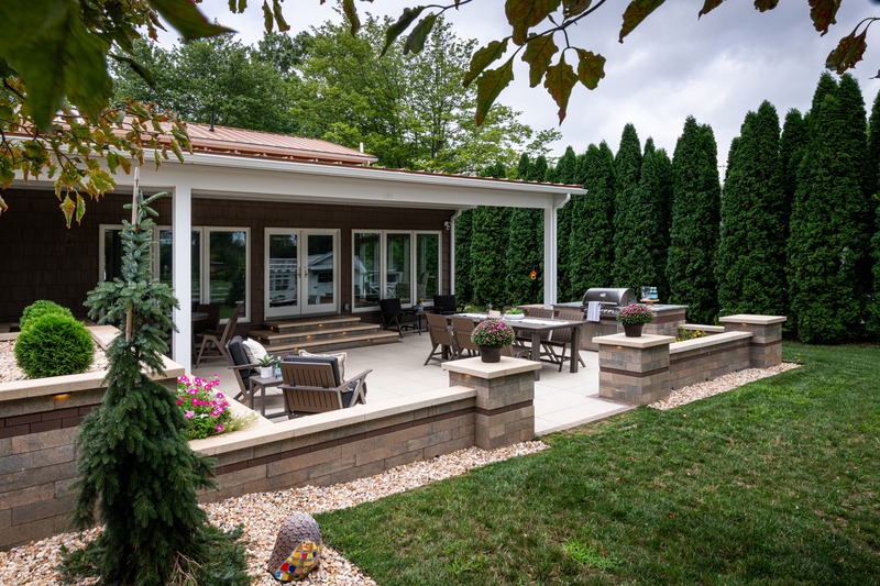 Cozy backyard patio with seating, dining table, and grill. Surrounded by lush greenery, flowers, and stone walls under a covered porch.