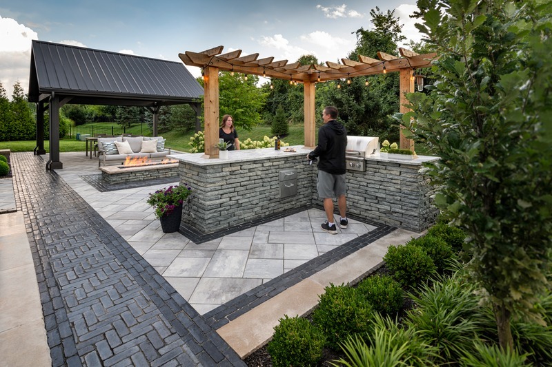 Two people in a stylish outdoor kitchen with stone counters, wooden pergola, and nearby seating area. Greenery and paved paths surround them.