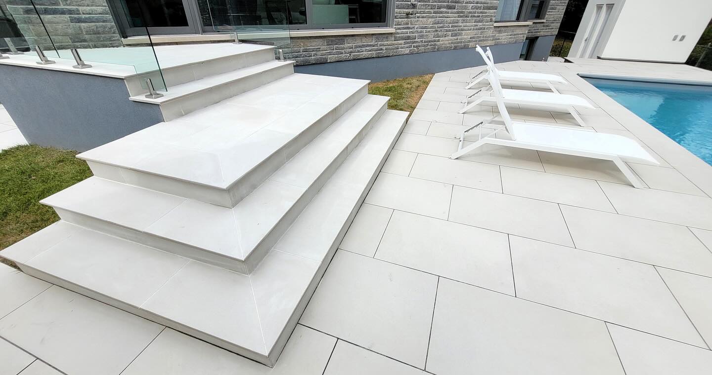 Modern patio with white tiled steps, glass railing, and poolside loungers beside a pool. Contemporary building visible in the background.