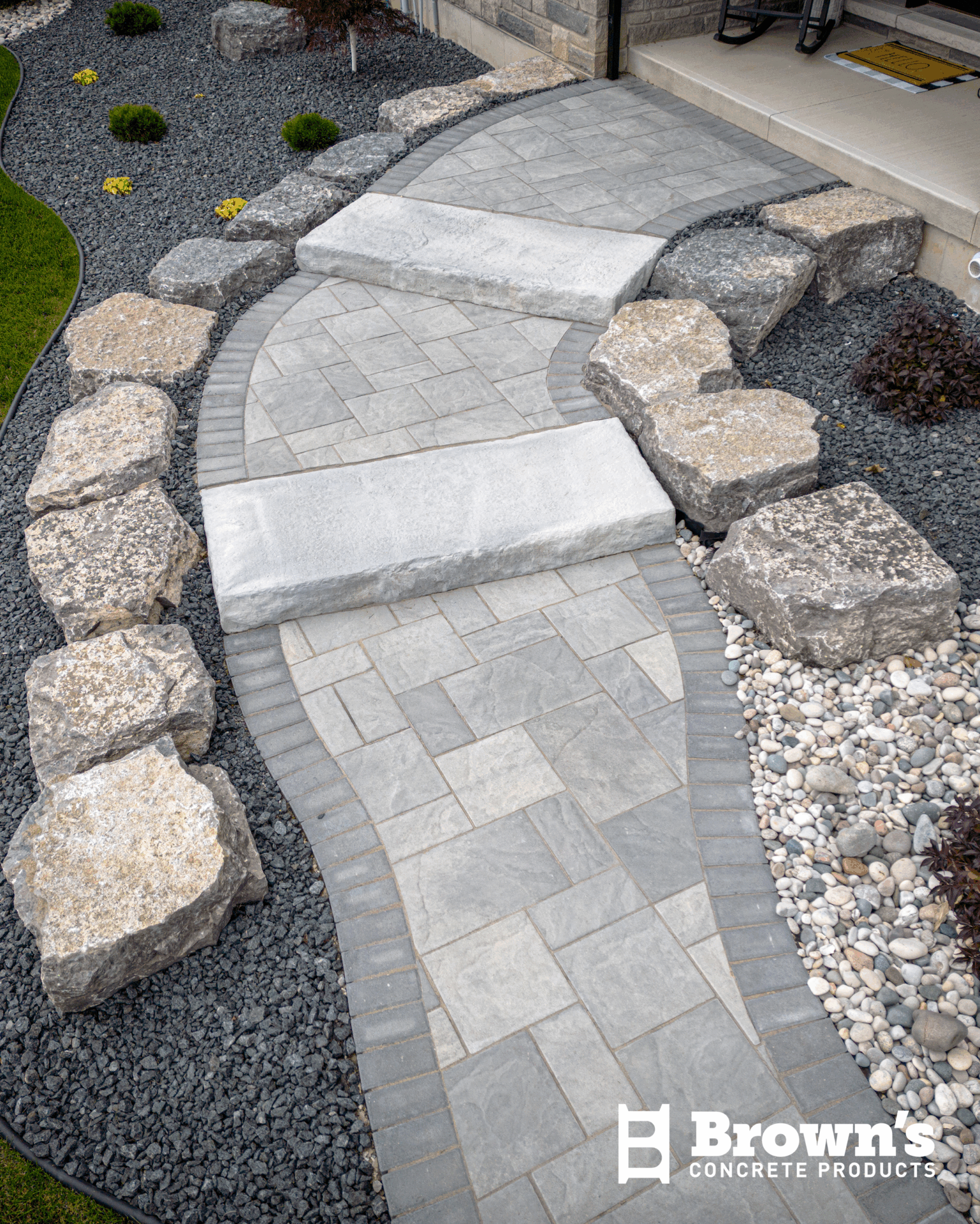 Curved stone walkway with gray pavers, surrounded by large rocks and small plants, leads up to a patio.