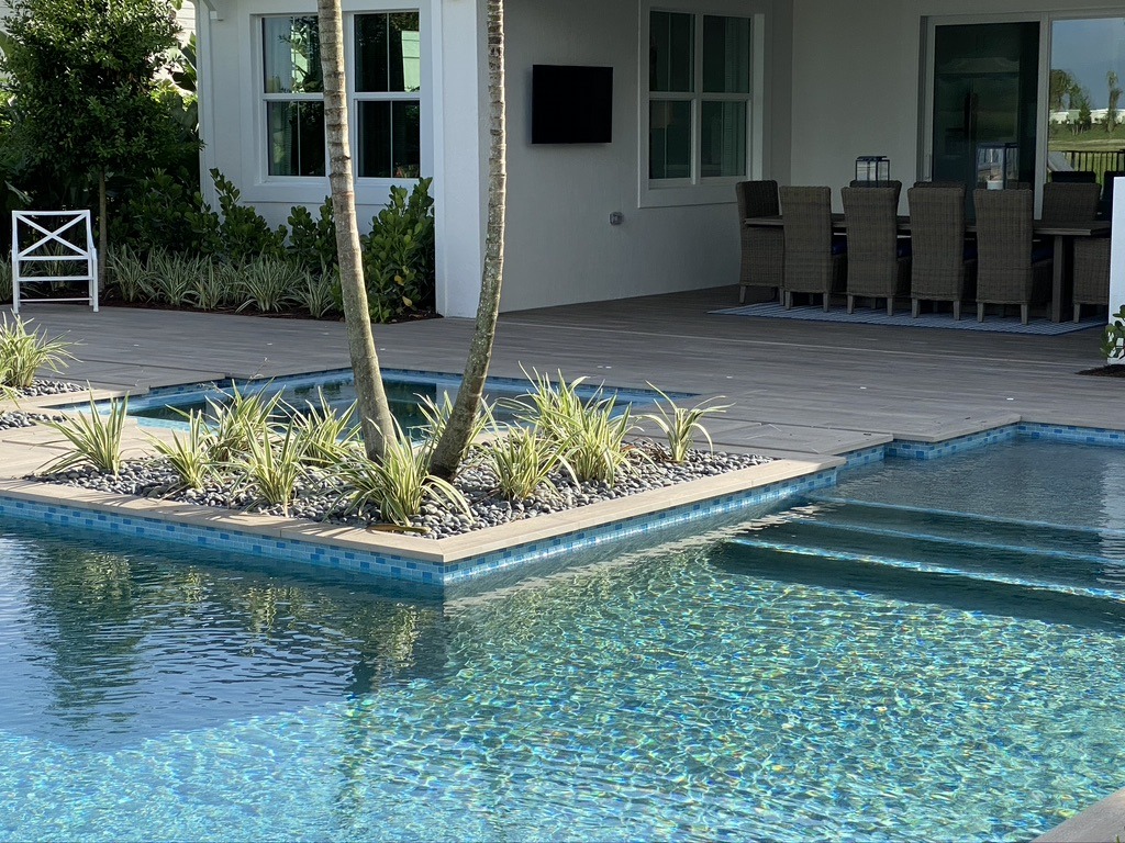 Modern outdoor area with a pool, wooden deck, plants, dining table, chairs, and a wall-mounted TV under a covered patio. Serene atmosphere.