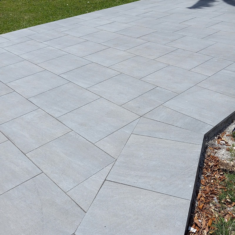 A clean, newly tiled patio with light grey square tiles bordered by green grass and a small mulch section on a sunny day.