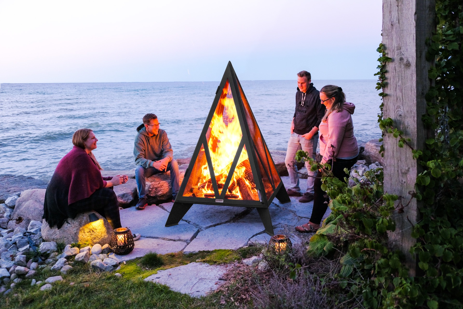Four people sit and stand around a triangular fire pit by the sea, surrounded by rocks and greenery, during sunset.