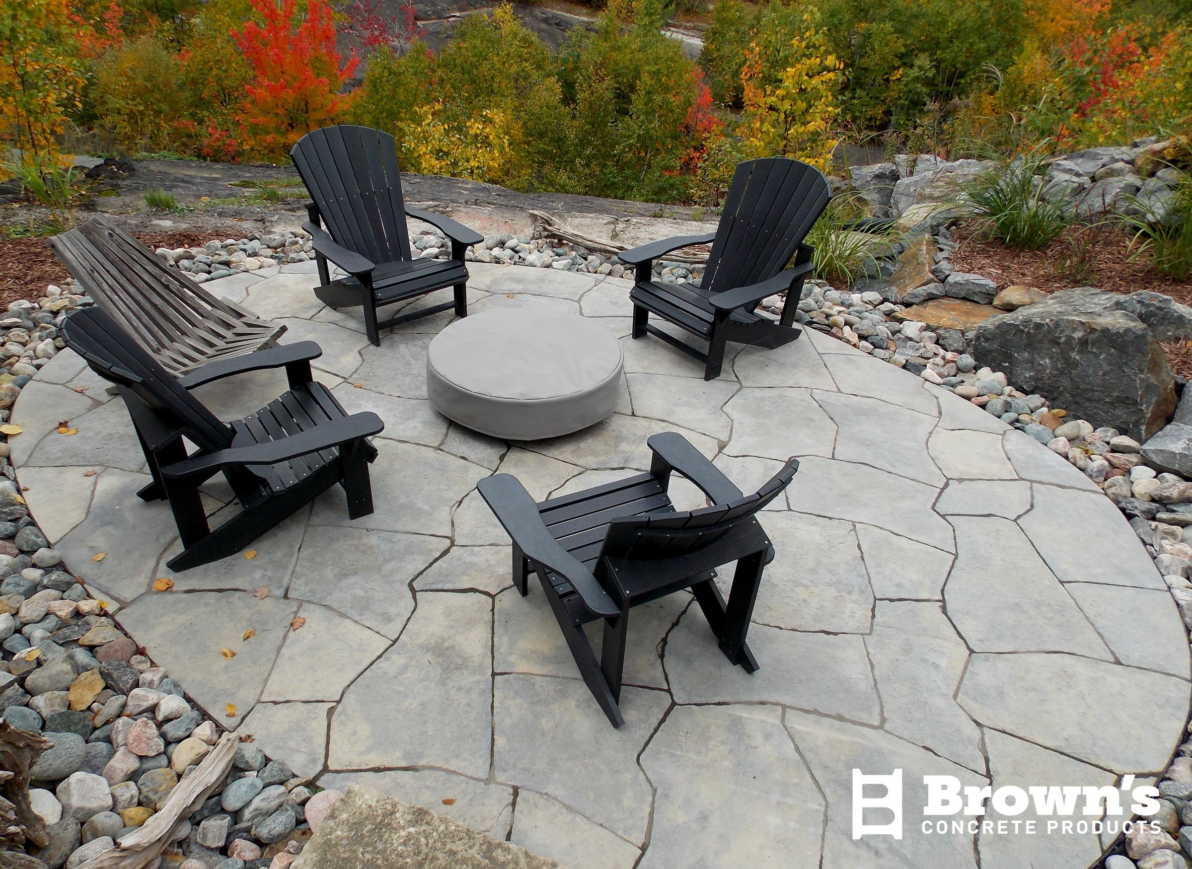 A patio features Adirondack chairs around a circular stone fire pit, surrounded by colorful autumn trees and rocky landscaping.