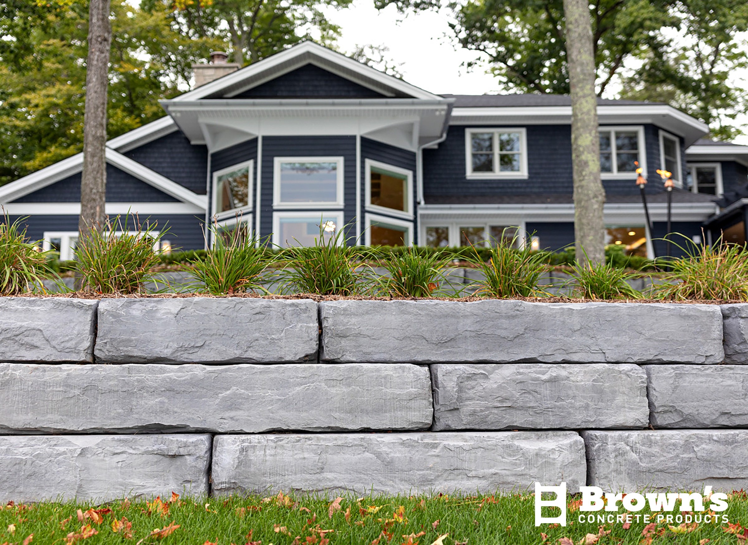 A modern house behind a stone retaining wall with greenery and trees, under a cloudy sky. Label: Brown's Concrete Products.