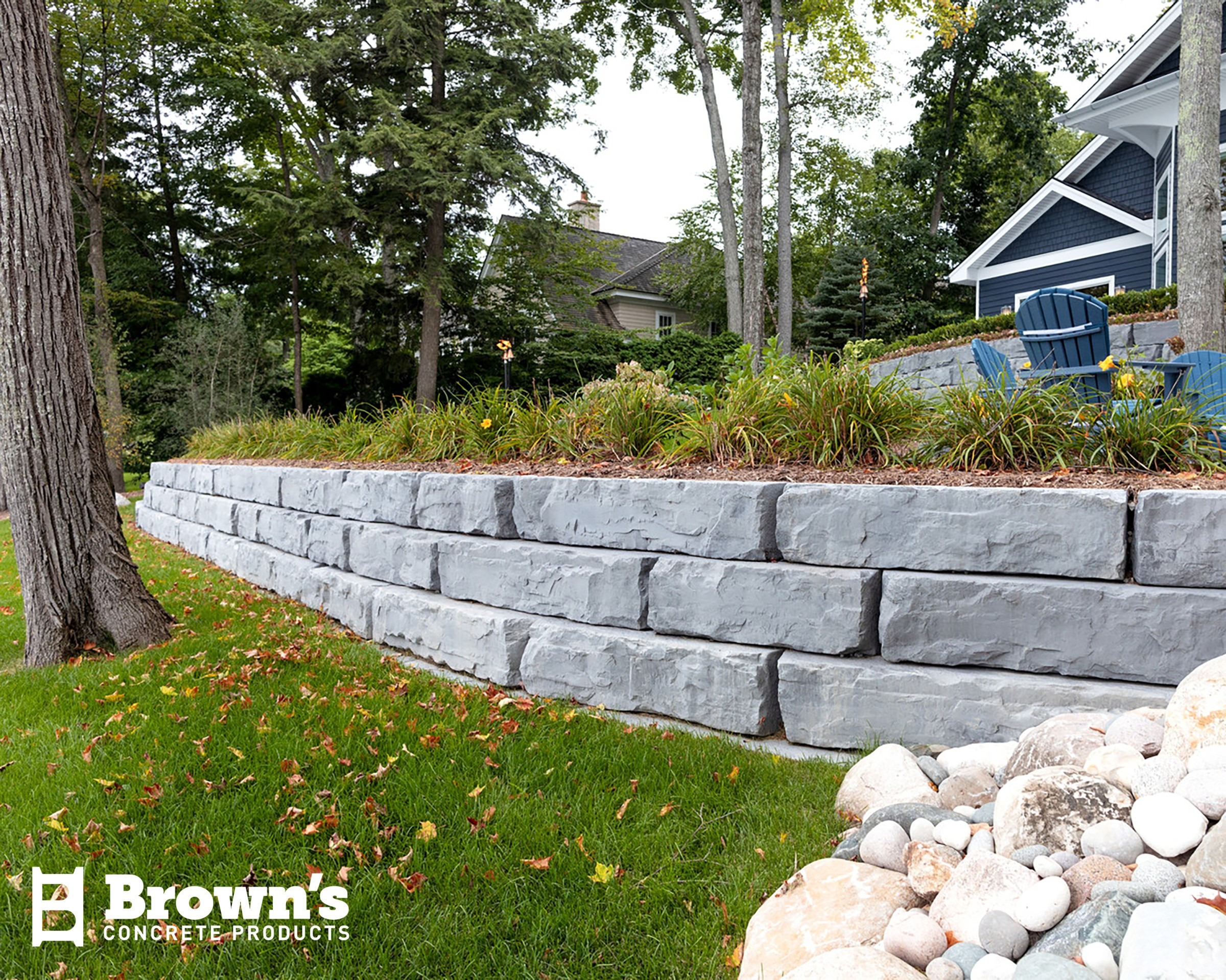 A tiered stone retaining wall with greenery and blue chairs. Trees surround the area. Lawn and decorative rocks in the foreground.