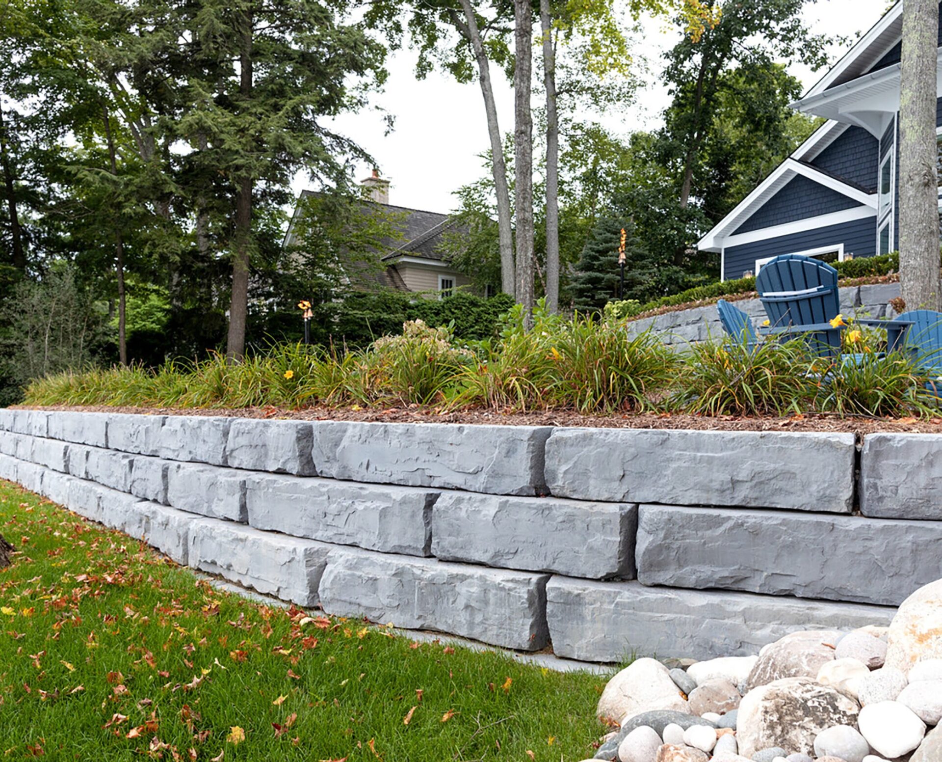 Terraced garden features large stone retaining wall, lush greenery, trees, blue house, and two blue chairs, providing a peaceful outdoor setting.