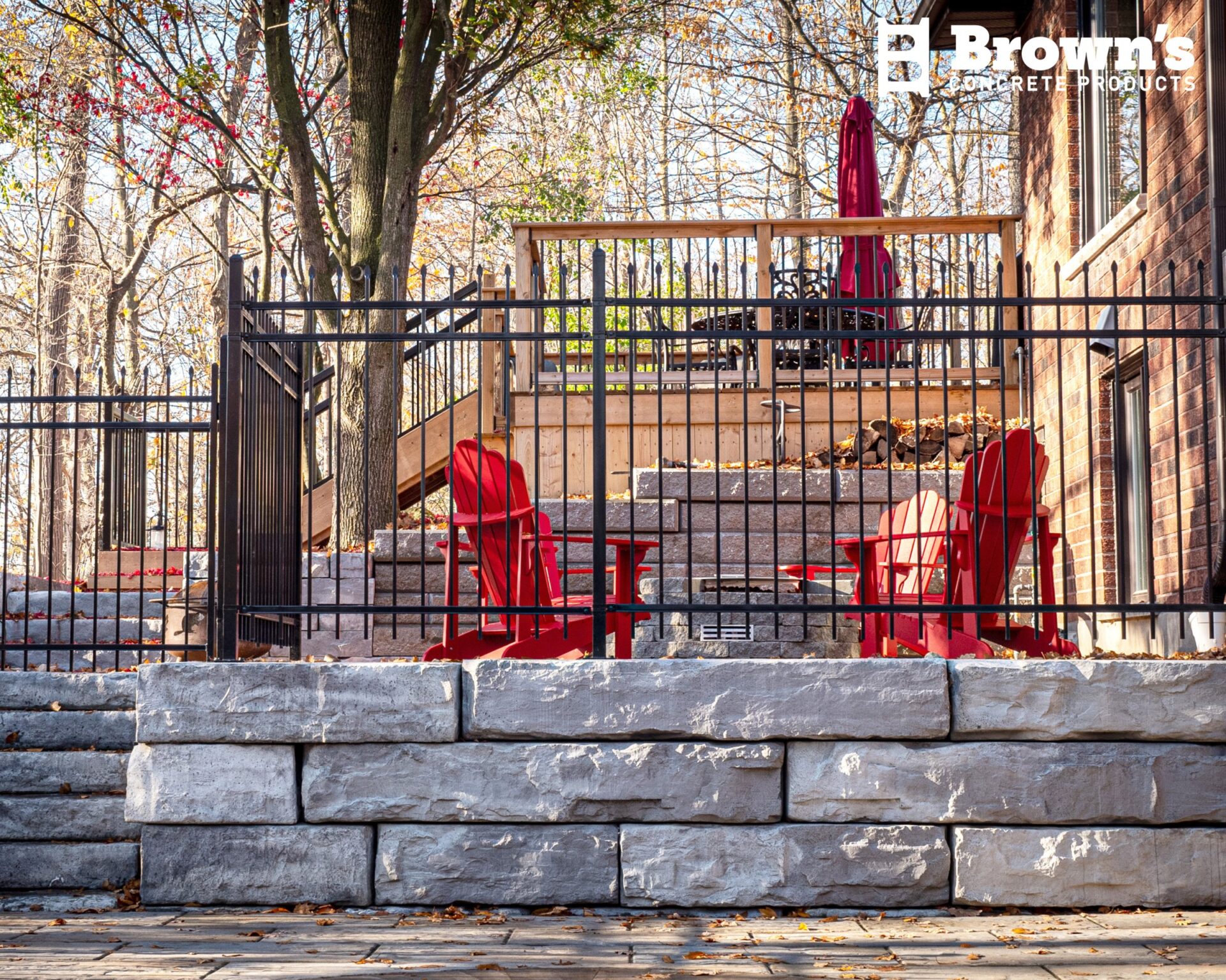 A serene outdoor patio with red chairs, surrounded by trees and a metal fence. Stone walls add rustic charm to the setting.
