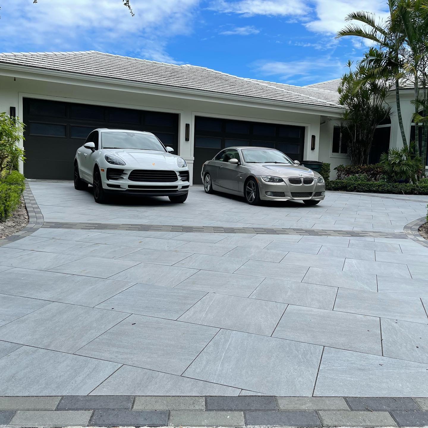 A modern house with a stone driveway features two luxury cars parked, surrounded by greenery, under a partly cloudy blue sky.