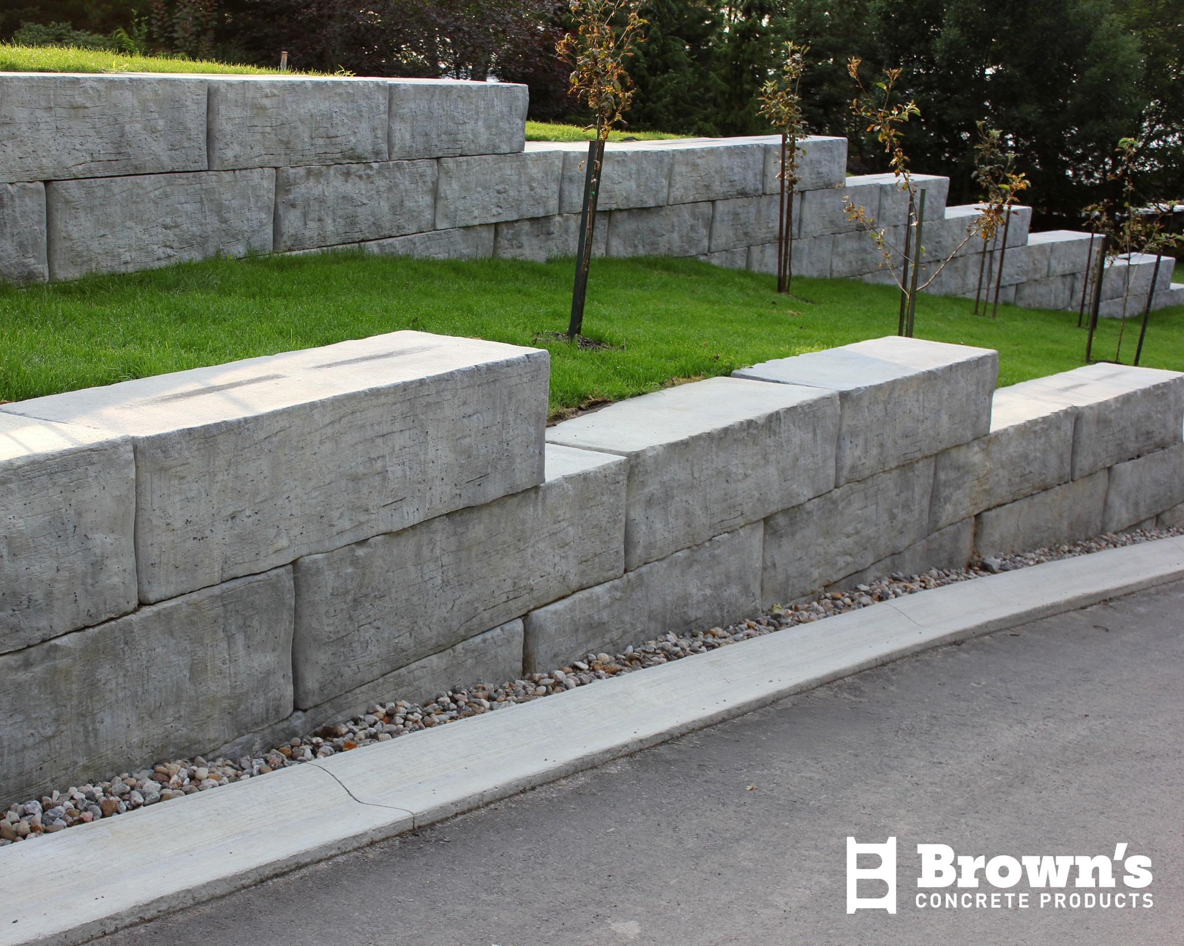 Tiered concrete retaining walls with young trees, lush green grass, and a pebble-lined edge on a sunny day. Brown's Concrete Products.