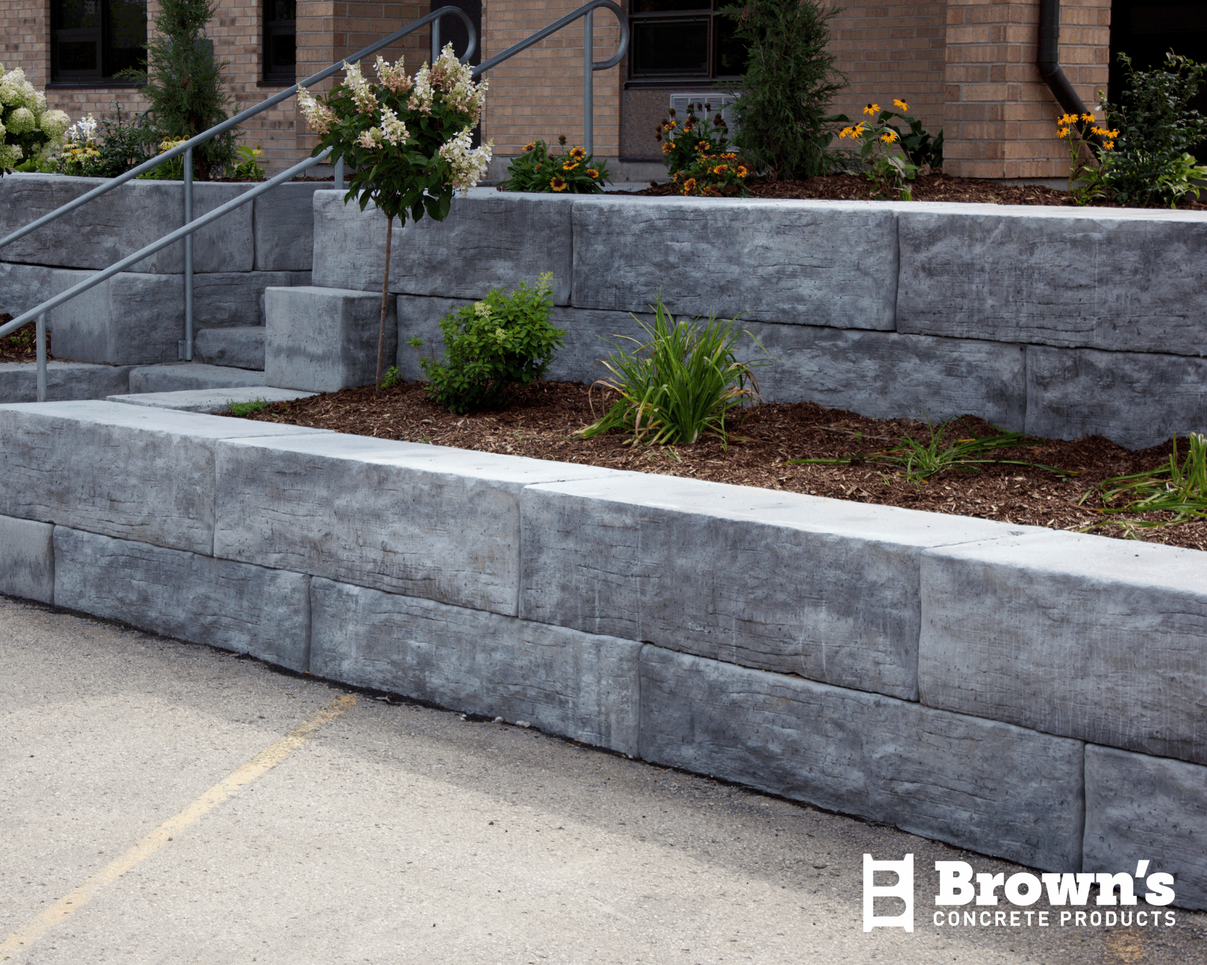 A concrete retaining wall with metal railings, surrounded by plants and flowers, is situated outside a brick building. No people are present.