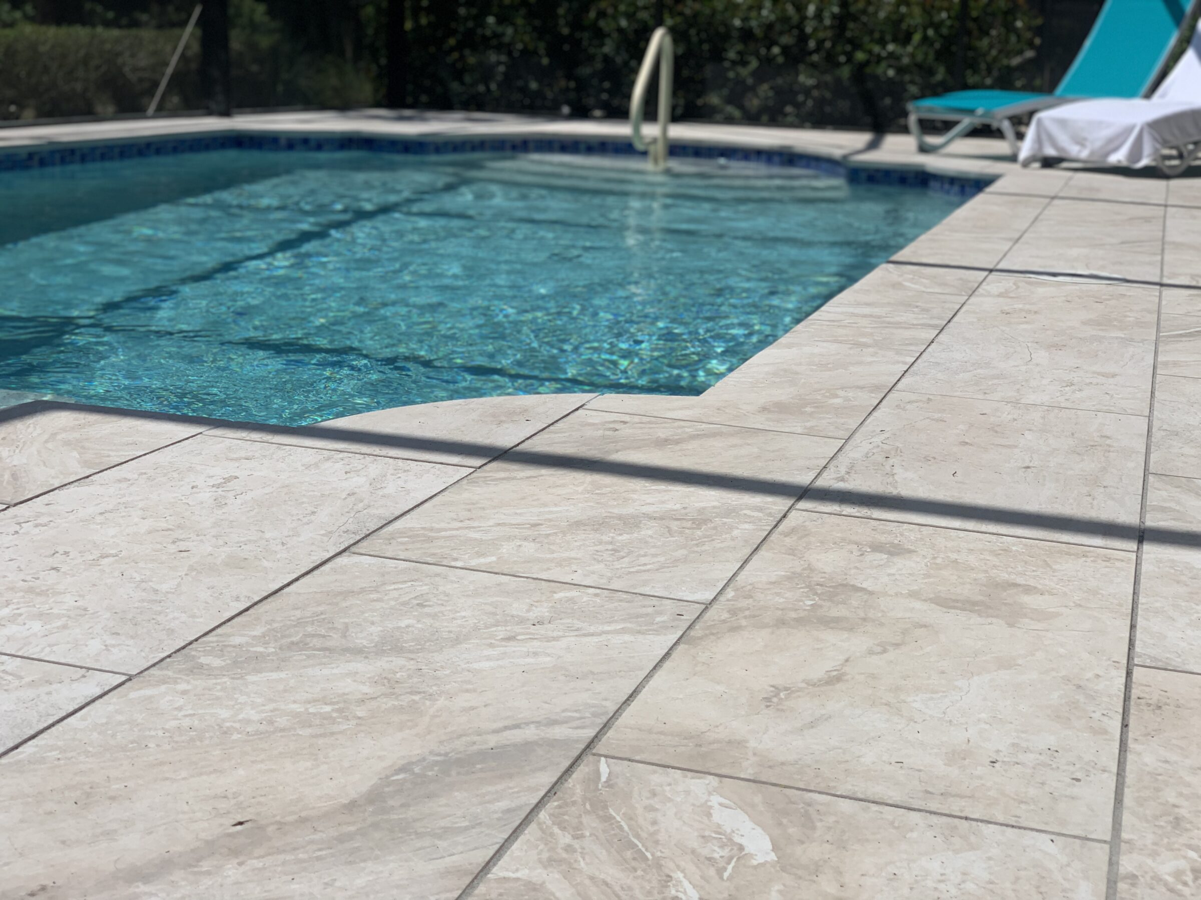 A serene outdoor pool with clear water, lounge chair, light-colored tiled deck, and natural surroundings, reflecting a sunny, relaxed atmosphere.