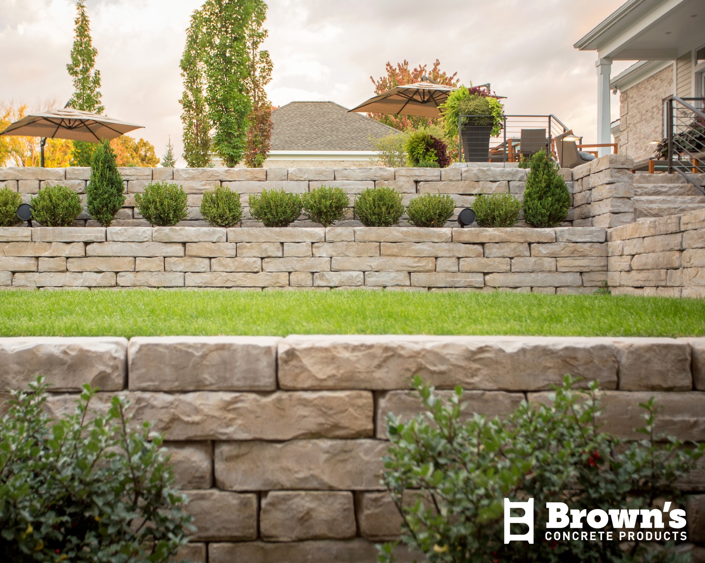 Tiered stone garden with lush green grass, shrubs, and patio furniture under umbrellas. Brown's Concrete Products logo visible in corner.