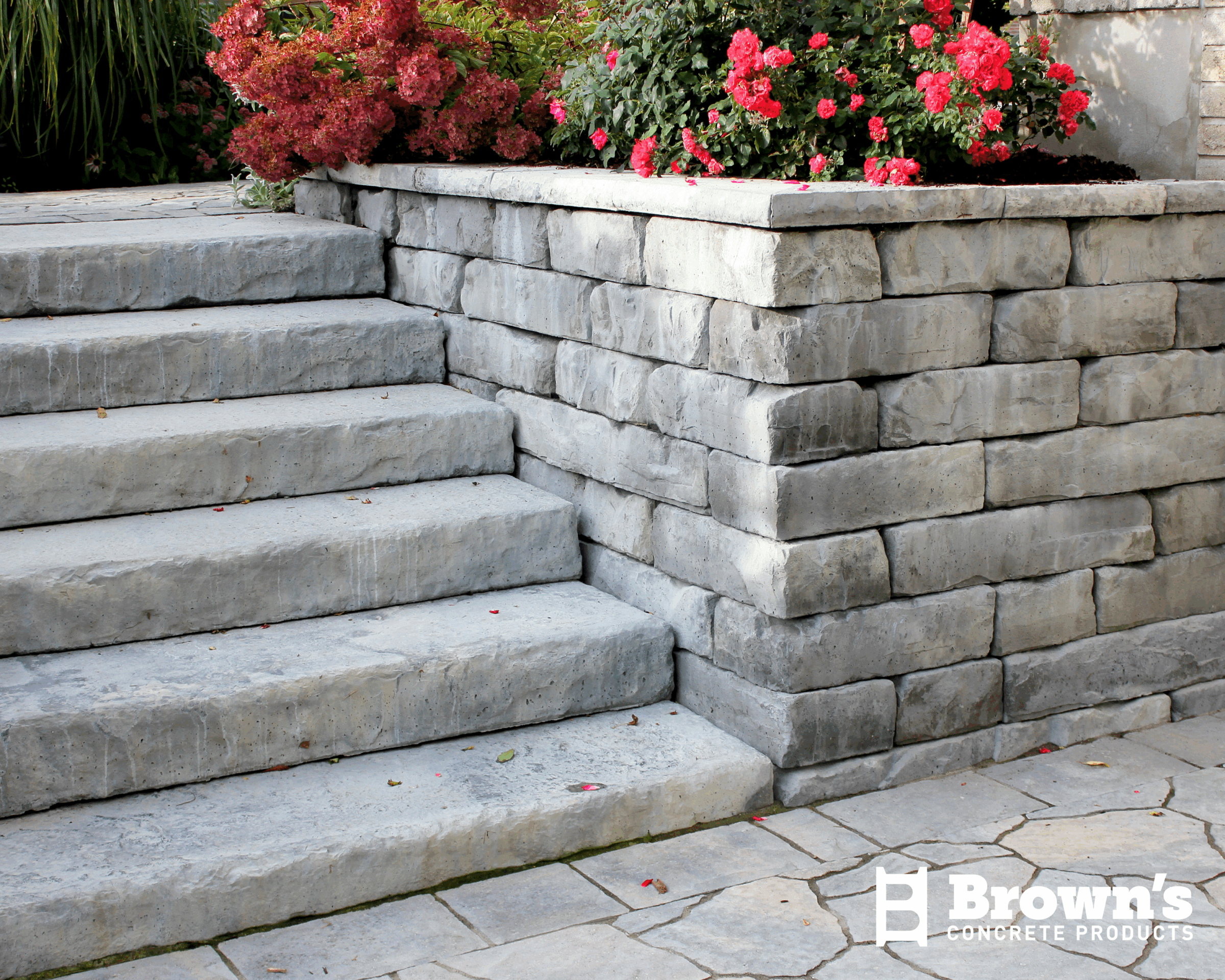 Stone steps lead to a garden bed with vibrant red flowers. The setting includes a concrete block retaining wall and paved stones.