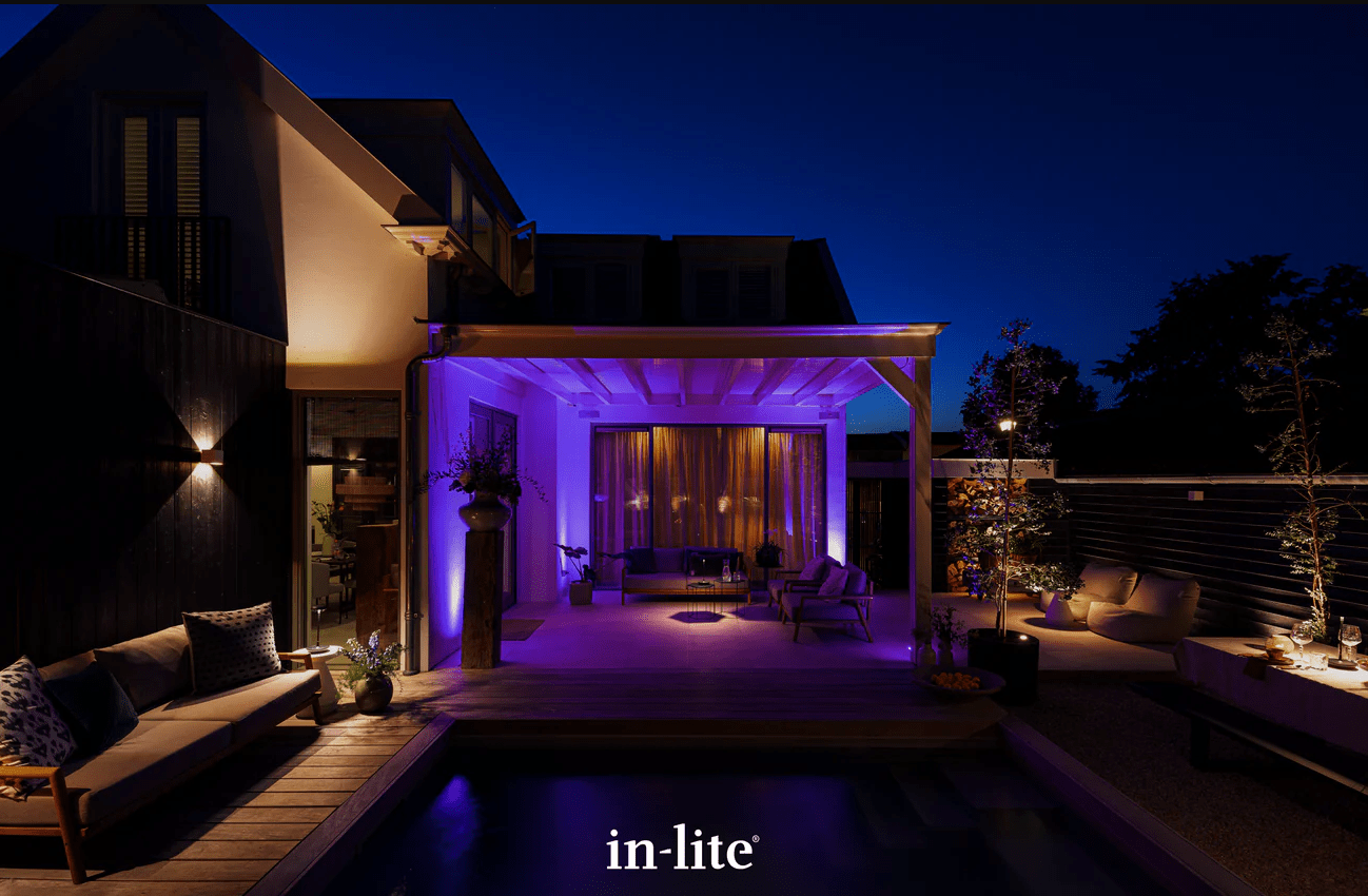 Modern outdoor patio illuminated with purple and yellow lighting at night. Cozy seating area with plants and swimming pool beside the house.