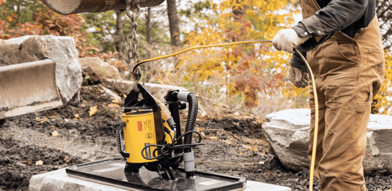 A person operates a construction tool, hooking a device onto a lifting mechanism. Autumn leaves and rocks surround the work area.