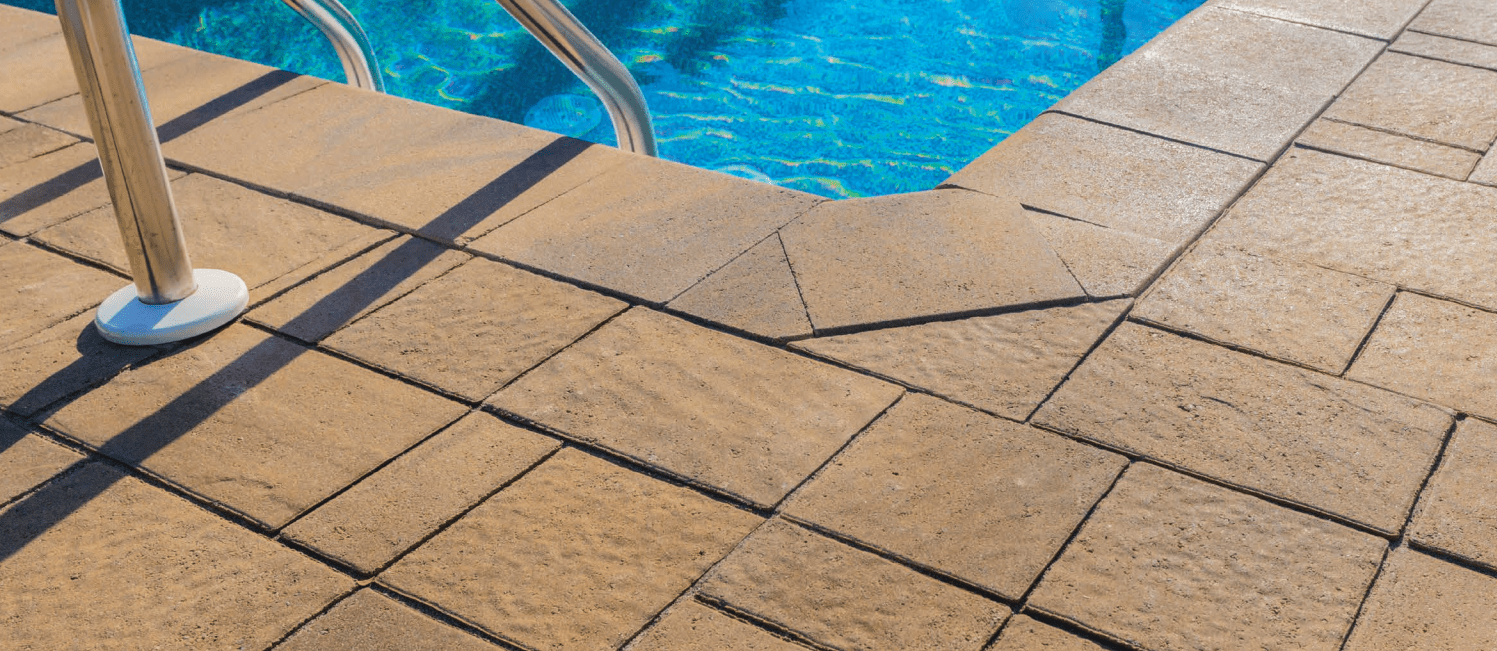 Close-up of a sunny poolside with blue water, metal handrail, and textured square paving stones casting shadows.