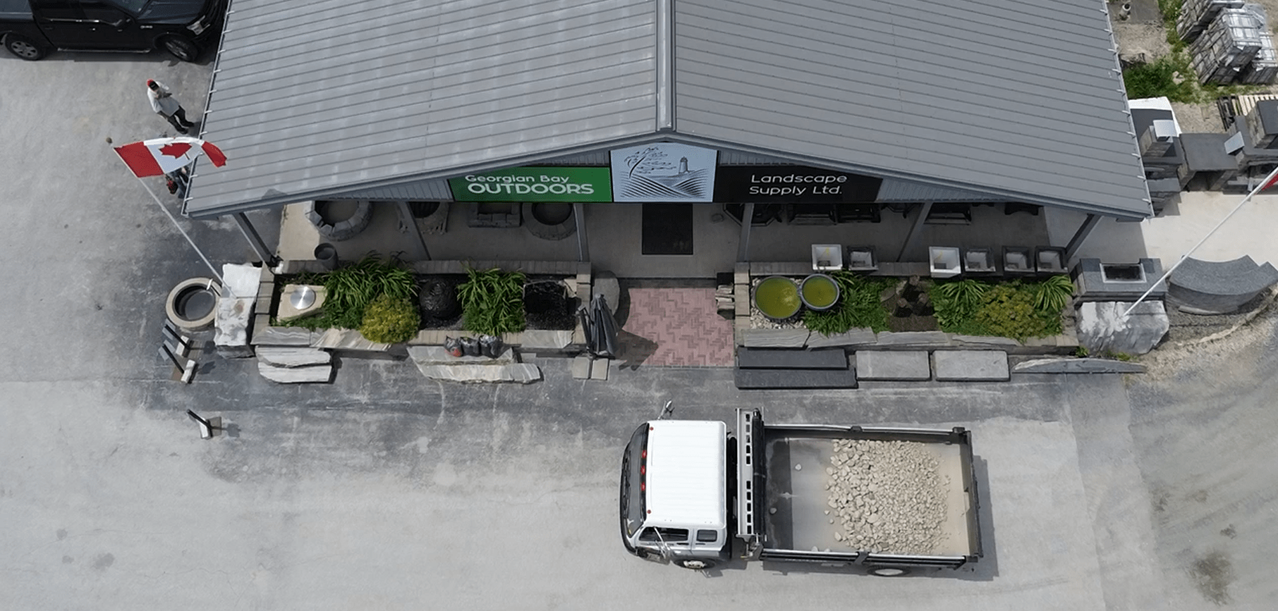 Aerial view of a landscape supply store with a Canadian flag, delivery truck, and person nearby. Building features plants and stone displays.