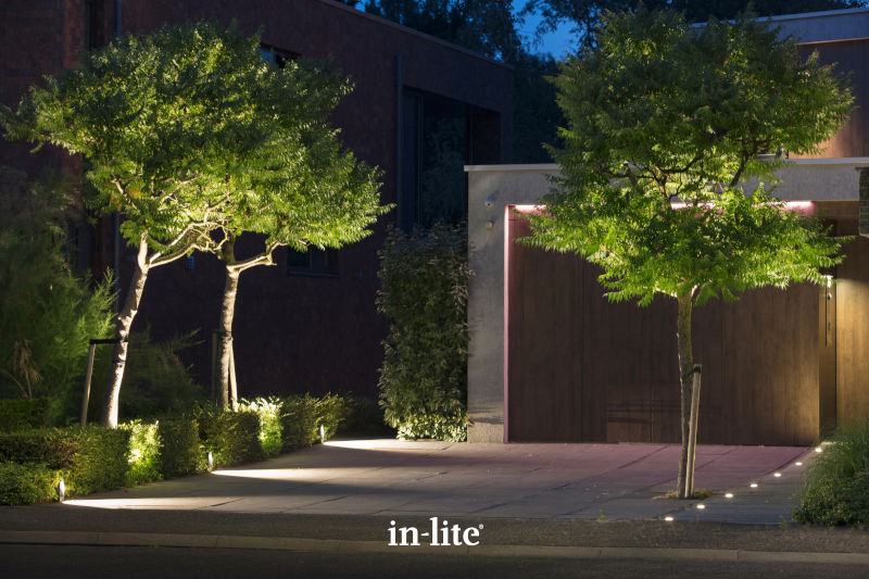 Modern house entrance illuminated by outdoor lighting, featuring two trees and neat landscaping, creating an inviting ambiance at dusk.