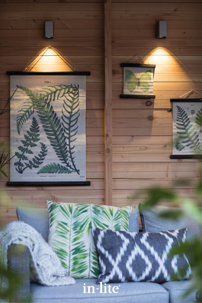 Wooden wall with fern and butterfly prints, illuminated by wall lights. Sofa features botanical-themed pillows, creating a cozy, nature-inspired setting.