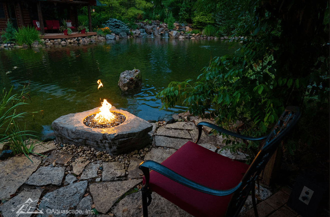 A cozy outdoor setting features a stone fire pit beside a tranquil pond with lush greenery and a comfortable red-cushioned chair.