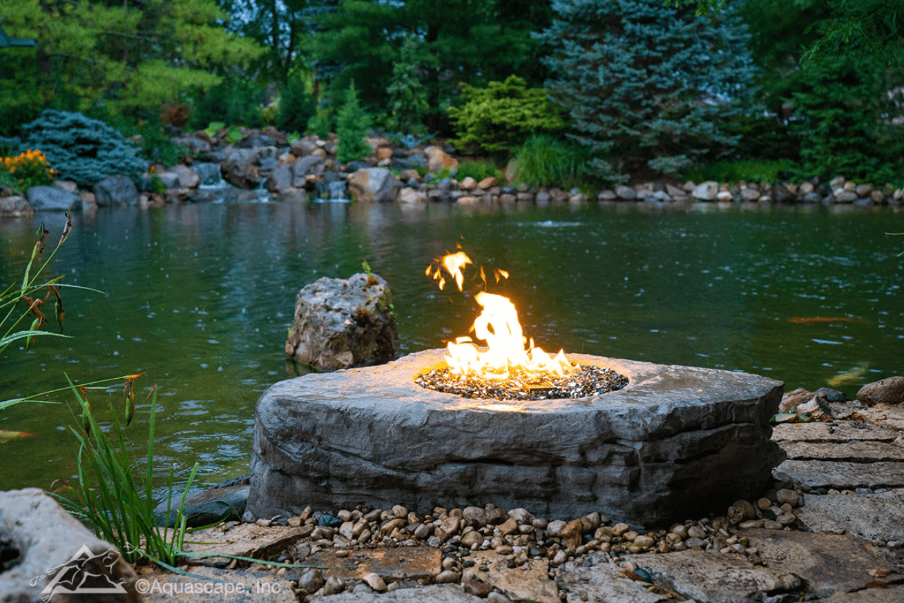 A cozy outdoor scene with a stone fire pit, surrounded by lush greenery and a serene pond, creating a tranquil ambiance.