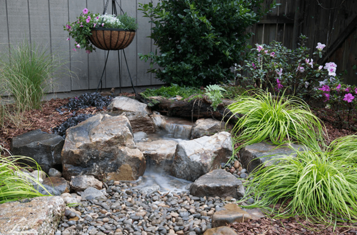 A tranquil garden with a small waterfall, surrounded by lush greenery, colorful flowers, rocks, and a hanging basket.