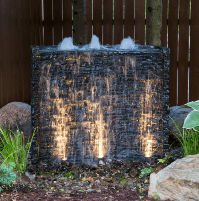 A garden water feature with illuminated cascading water, surrounded by stone and greenery, creates a serene and calming atmosphere in an outdoor setting.