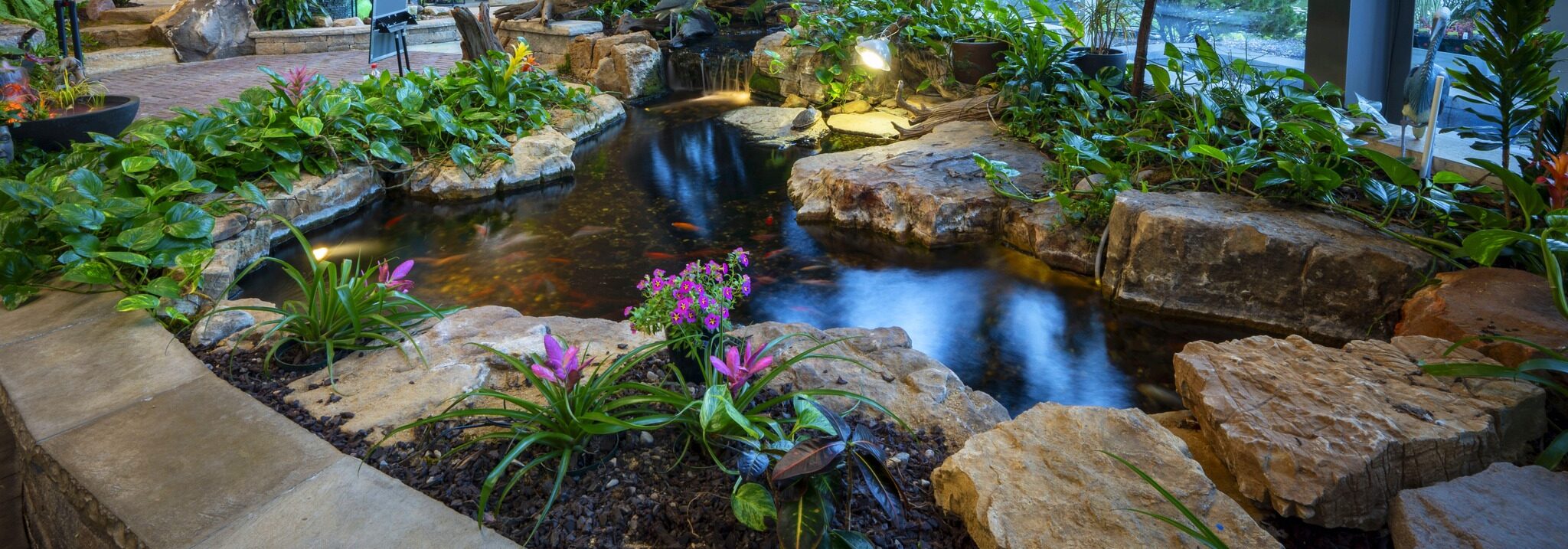 Indoor garden with lush greenery, vibrant flowers, and a small waterfall into a koi pond. Rocks and plants surround the tranquil water feature.
