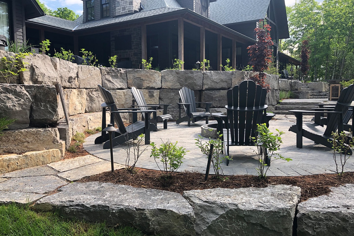 Stone patio with Adirondack chairs, surrounded by large stone walls and greenery. Modern house with wooden elements in the background.