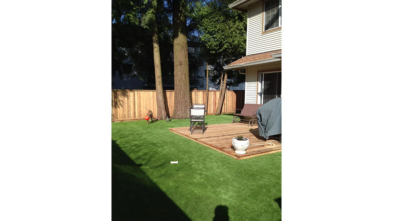A backyard with artificial turf, wooden deck, sitting area, and potted plant. A dog plays with a red ball near the fence.