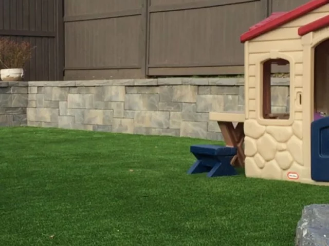 A backyard setting with artificial grass, a playhouse, and a stone retaining wall. A planter sits atop the wall, near a wooden fence.
