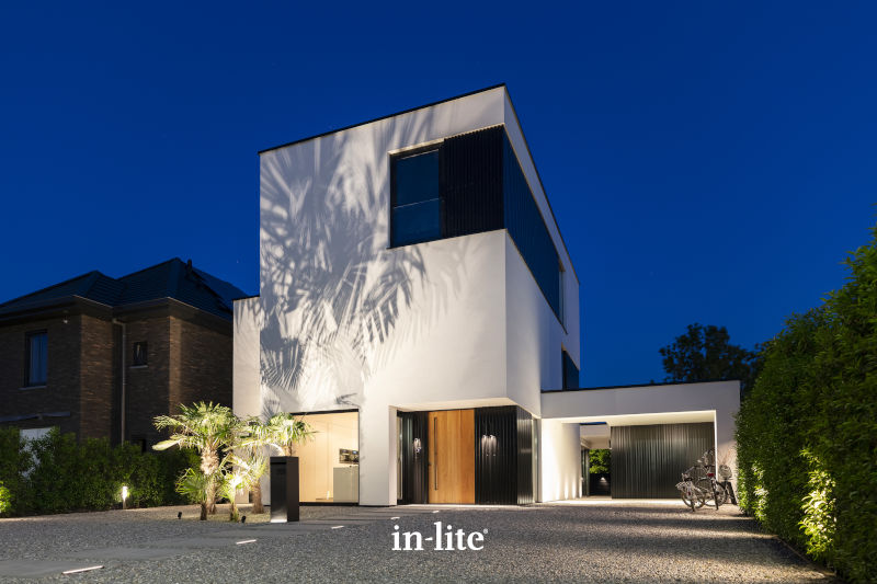Modern, two-story house with sleek design features illuminated at night, surrounded by trees and shrubbery. A bicycle is parked outside.