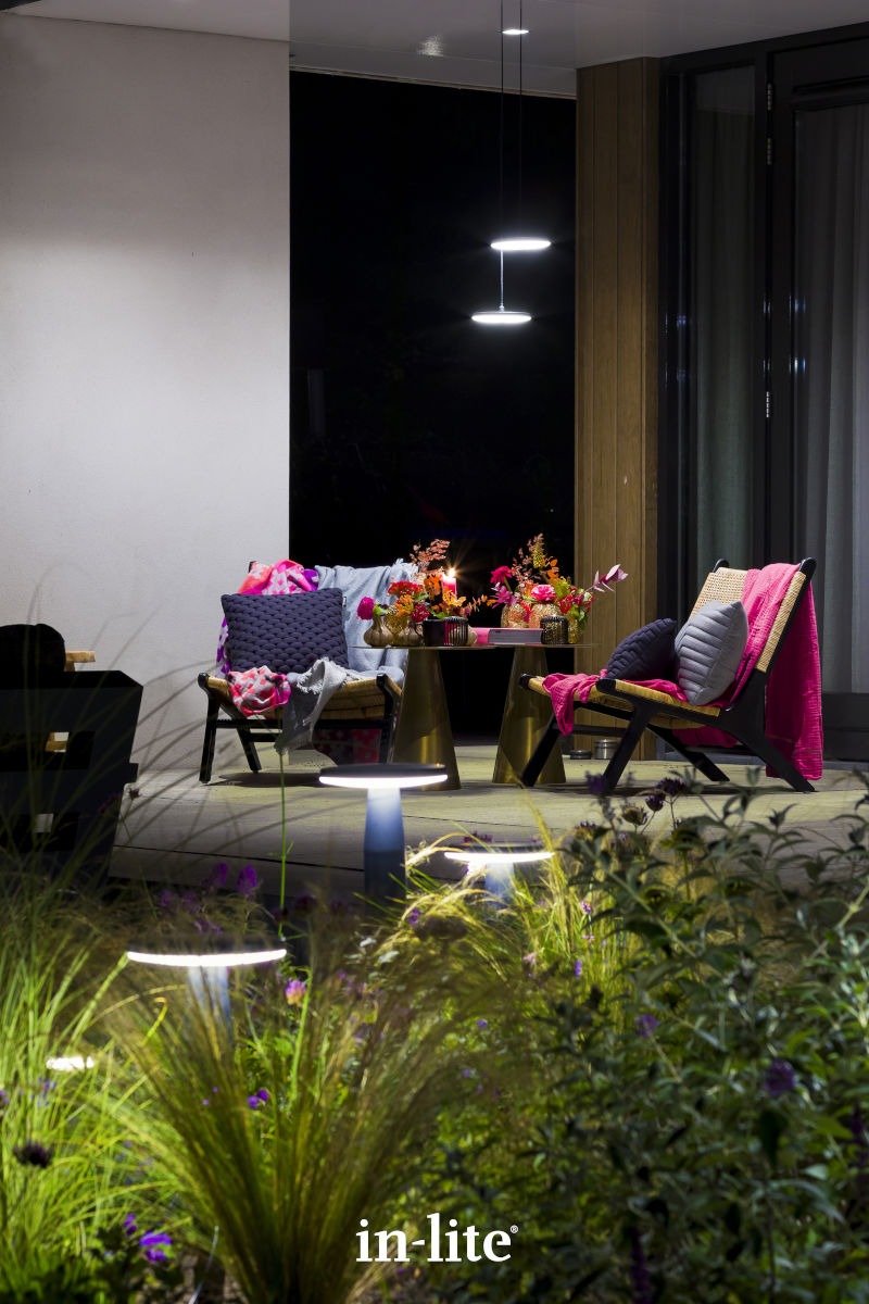 Cozy outdoor seating area with colorful cushions and decorative lighting, surrounded by plants. Modern chairs and table under soft evening illumination.