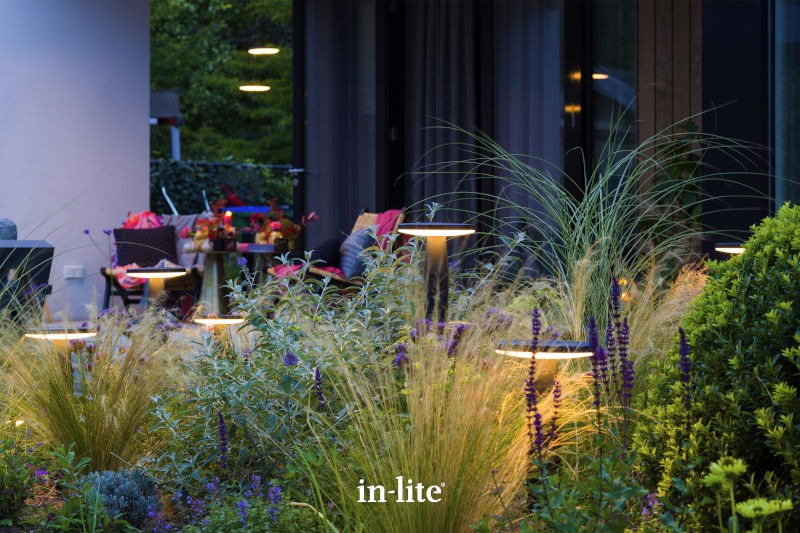 A cozy patio with modern outdoor lighting, surrounded by lush greenery, flowers, and comfortable seating in a tranquil garden setting.