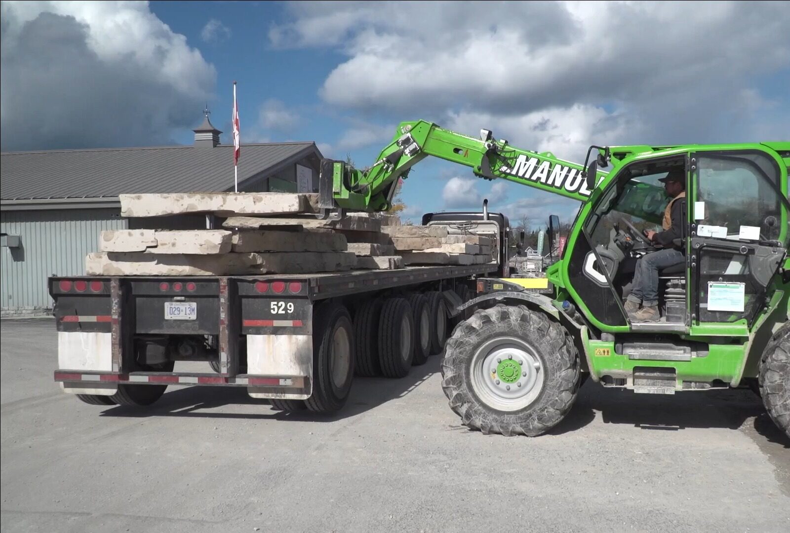 A person operates a green telehandler to load stone slabs onto a truck. A building with a Canadian flag is nearby.