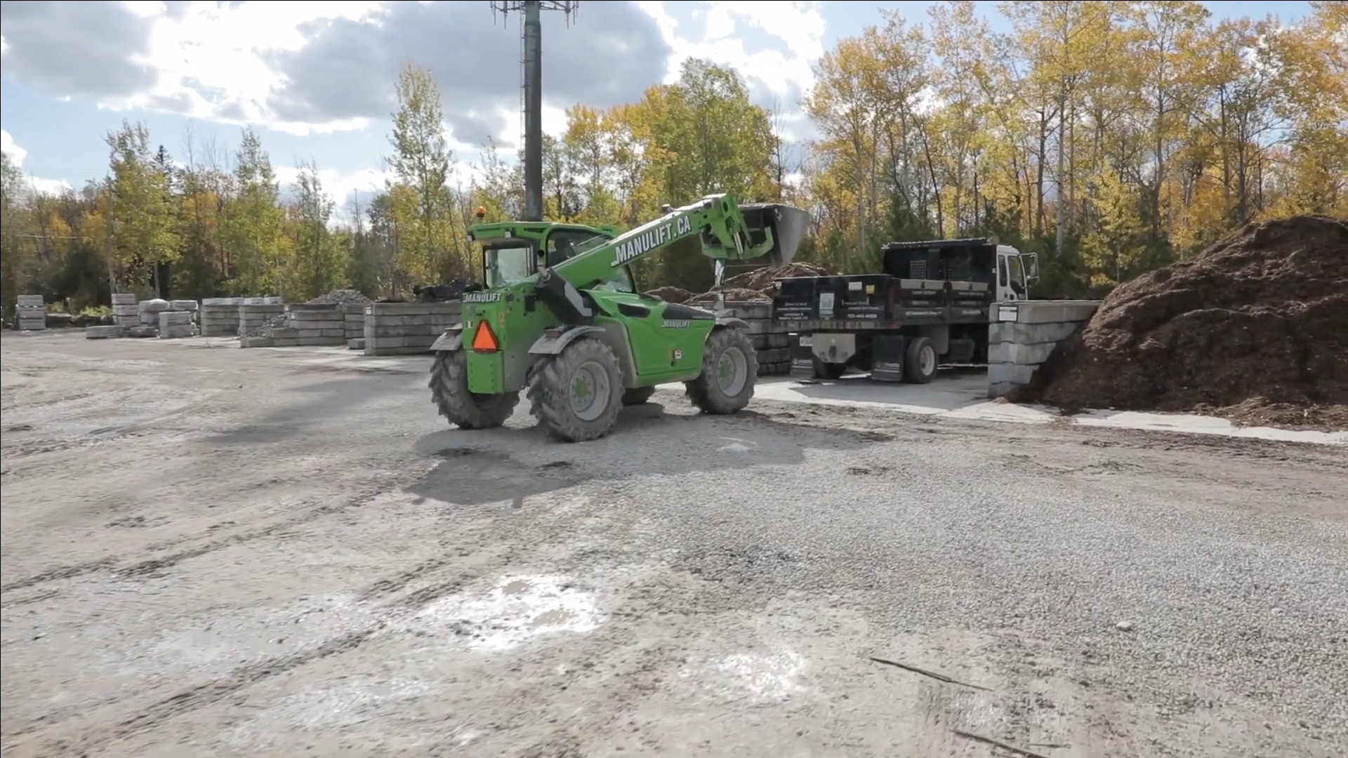 A green telehandler operates in a construction yard with autumn trees, concrete slabs, and a truck. Cloudy sky overhead.