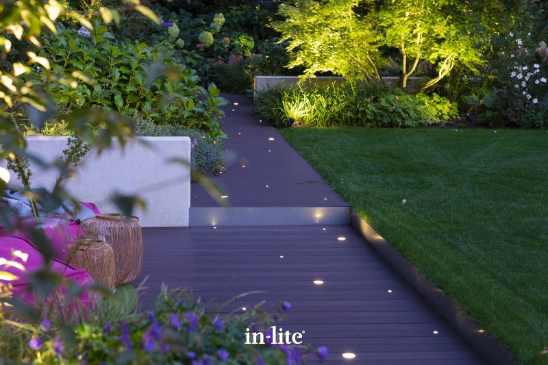 A serene garden features illuminated wooden pathways, lush greenery, and ambient lighting, creating an inviting and tranquil outdoor space for relaxation.