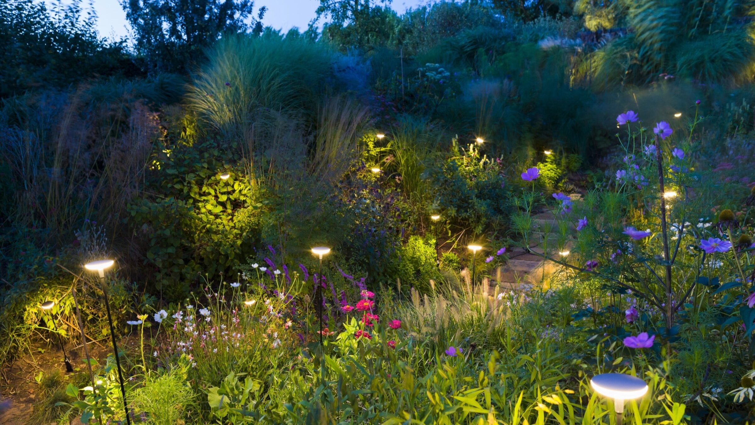 A garden at dusk, featuring vibrant flowers and foliage illuminated by soft, warm garden lights. No visible landmarks or historical buildings present.