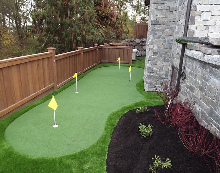A backyard putting green with three flags, bordered by a stone wall and wooden fence, surrounded by plants and trees.