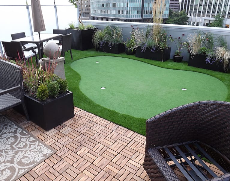 Rooftop terrace with a small putting green, surrounded by planters and seating. Cityscape background with modern buildings visible in the distance.