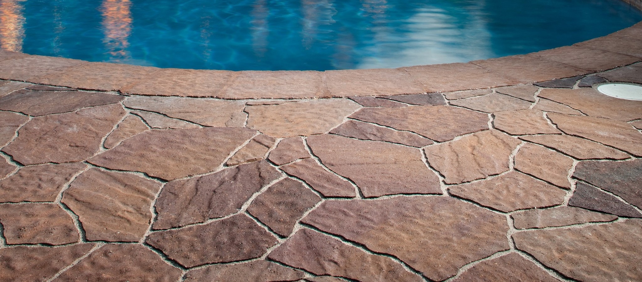 Stone tiles form a decorative patio around a clear blue swimming pool, creating an inviting and tranquil outdoor setting.