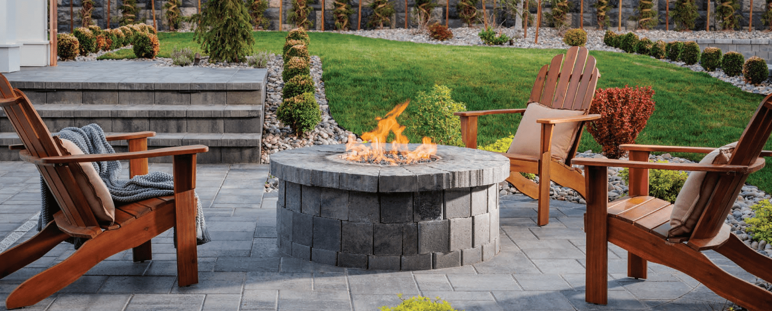 A cozy backyard patio features a stone fire pit surrounded by wooden chairs, lush green grass, and manicured bushes for a serene ambiance.