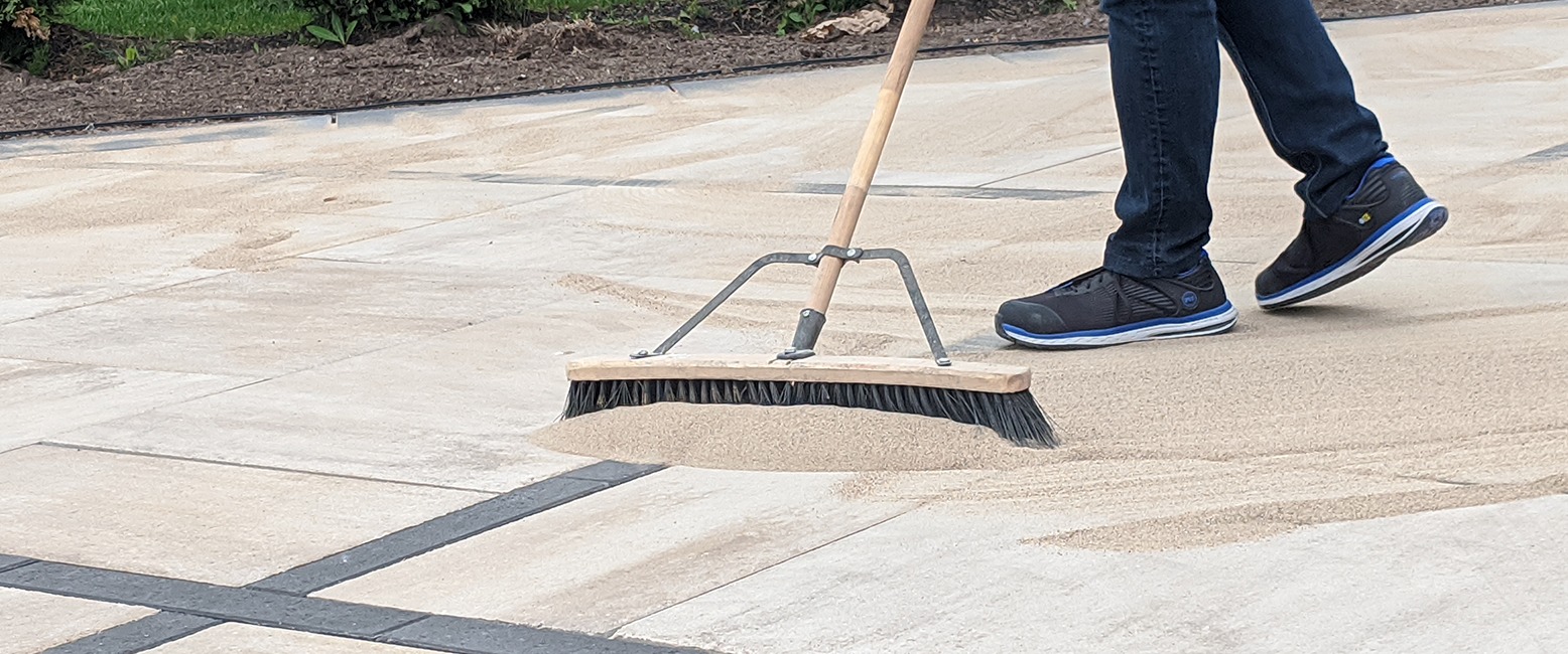 A person spreads sand on paving slabs with a large broom, wearing jeans and sneakers. Background shows grass and soil.
